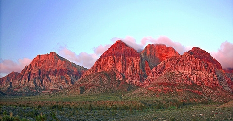 Sléibhte dearga Red Rock Canyon faoi sholas te, le scamaill agus spéir ghorm os a gcionn.
